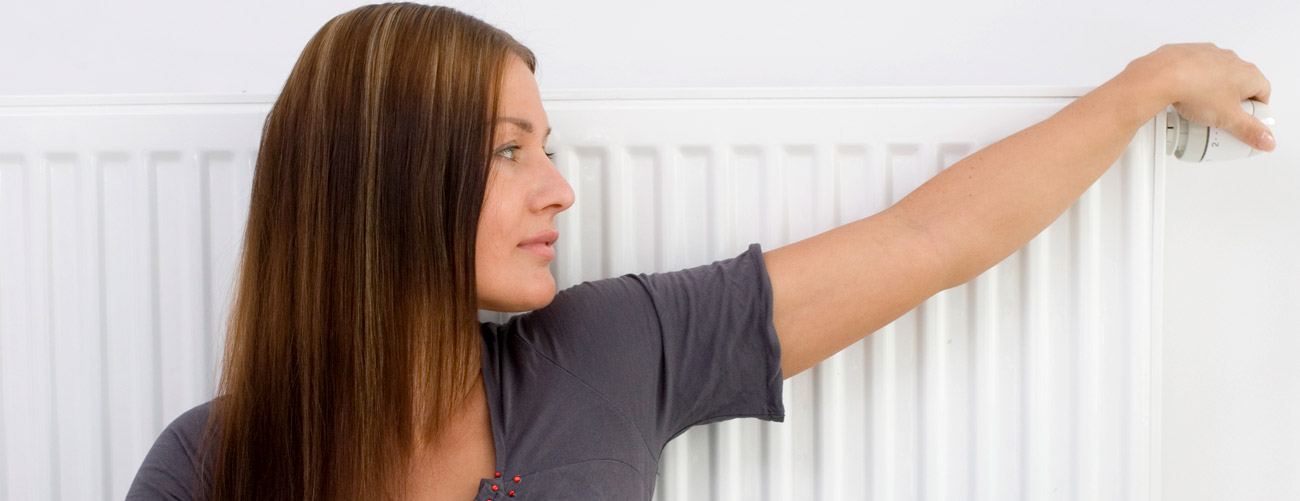 Frau lehnt sich sitzend an Heizung und dreht am Thermostat